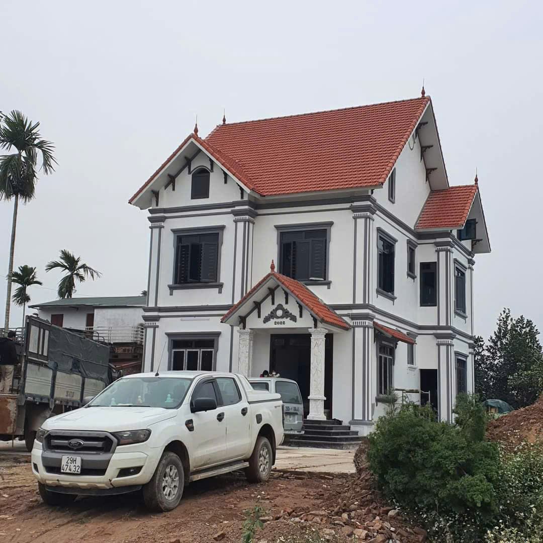 Nhà hai tầng mái ngói đỏ, tường trắng viền xám, xe bán tải đậu trên nền đất trước nhà.