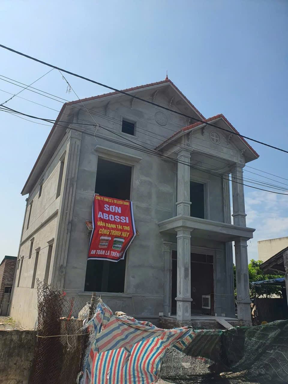 Nhà dân dụng mái đỏ đang hoàn thiện, băng rôn treo trên cửa sổ và bạt che vật tư trước nhà.