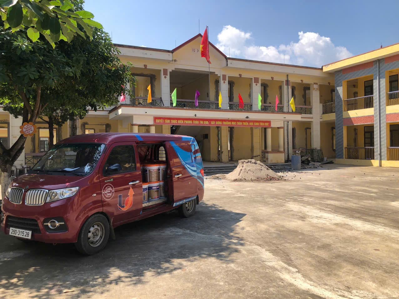 Xe tải nhỏ mở cửa bên, thùng sơn xếp trong khoang, đậu trong sân trường học đang sửa chữa.